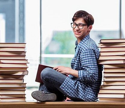 Young man with glasses sits between 2 stacks of books on a table