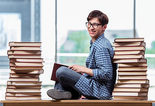 Junger Mann mit Brille sitzt zwischen 2 Bücherstapeln auf einem Tisch