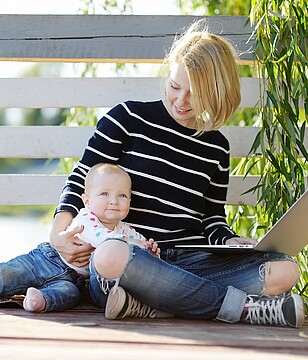 Junge Frau im Schneidersitz mit Baby im Arm und Laptop auf den Knien