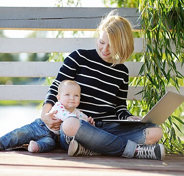 Junge Frau im Schneidersitz mit Baby im Arm und Laptop auf den Knien